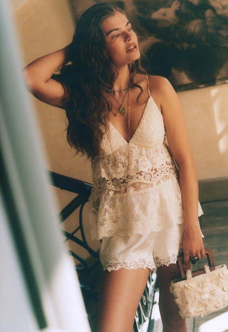 A woman wearing a cream lace camisole top and matching lace shorts, holding a small cream floral handbag, posed in a warm-toned indoor setting.