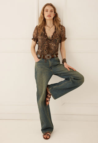 a-woman-modeling-a-brown-patterned-short-sleeve-blouse-and-wide-leg-blue-jeans-with-brown-sandals-posing-against-a-white-paneled-wall