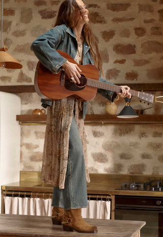 a-woman-stands-in-a-rustic-kitchen-setting-holding-and-playing-an-acoustic-guitar-she-is-wearing-a-blue-denim-jacket-blue-jeans-and-brown-leather-boots