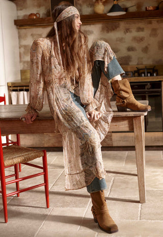 a-woman-in-a-bohemian-style-patchwork-long-sleeve-dress-and-blue-jeans-sits-on-a-wooden-table-in-a-rustic-kitchen-wearing-brown-suede-boots