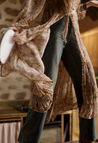 a-close-up-lifestyle-shot-of-a-person-wearing-a-long-flowing-beige-and-brown-patterned-sheer-cardigan-or-duster-over-dark-blue-jeans-holding-a-white-plate-in-a-rustic-indoor-setting
