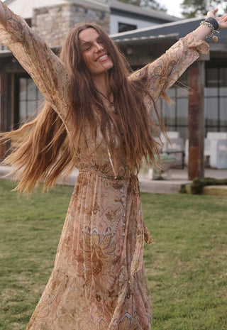 a-woman-with-long-brown-hair-wearing-a-flowing-beige-and-brown-patterned-bohemian-maxi-dress-posing-outdoors-with-her-arms-raised-in-front-of-a-modern-house-and-grassy-lawn