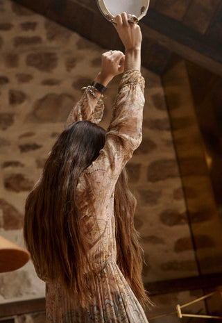 a-woman-with-long-dark-hair-seen-from-behind-wearing-a-beige-and-brown-patterned-long-sleeve-dress-reaching-upward-in-a-rustic-stone-walled-room