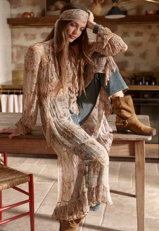 a-woman-in-a-bohemian-style-room-wearing-a-long-loose-beige-and-brown-patterned-tunic-dress-with-a-matching-headscarf-blue-jeans-and-tan-boots-sitting-on-a-wooden-table