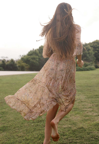 a-woman-with-long-brown-hair-runs-barefoot-across-a-grassy-field-viewed-from-behind-wearing-a-flowing-beige-bohemian-lace-dress-with-a-subtle-floral-pattern