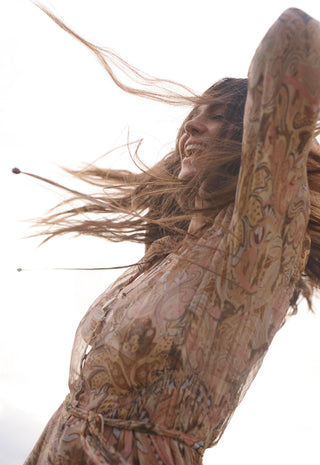a-woman-with-long-wind-blown-brown-hair-wearing-a-loose-patterned-beige-dress-with-brown-and-pink-paisley-like-motifs-captured-against-a-bright-white-sky