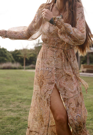 a-woman-outdoors-in-a-grassy-park-wearing-a-long-flowing-boho-style-maxi-dress-with-a-high-slit-the-dress-has-an-intricate-paisley-and-floral-pattern-in-muted-beige-tan-and-brown-tones-with-subtle-gold-and-blue-accents
