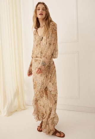 a-woman-modeling-a-long-sleeved-maxi-dress-with-a-beige-and-brown-paisley-pattern-standing-in-a-bright-minimalist-room-with-white-walls-and-a-cream-curtain