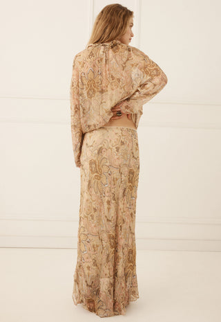 a-woman-stands-with-her-back-to-the-camera-wearing-a-matching-two-piece-set-consisting-of-a-long-sleeve-cropped-blouse-and-a-long-skirt-both-in-a-beige-paisley-pattern-with-brown-and-muted-gold-details