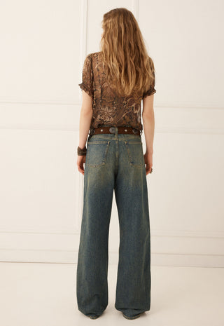 a-woman-viewed-from-behind-wearing-a-brown-patterned-short-sleeve-top-a-brown-belt-and-wide-leg-blue-jeans-against-a-white-paneled-wall