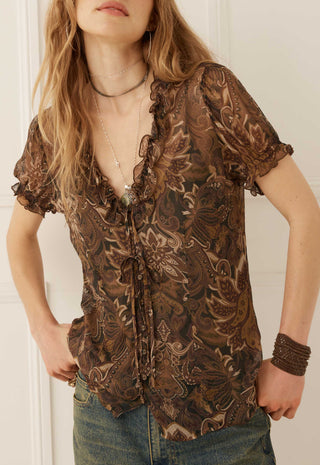a-woman-wearing-a-brown-and-beige-paisley-patterned-sheer-blouse-with-ruffle-trim-paired-with-blue-jeans
