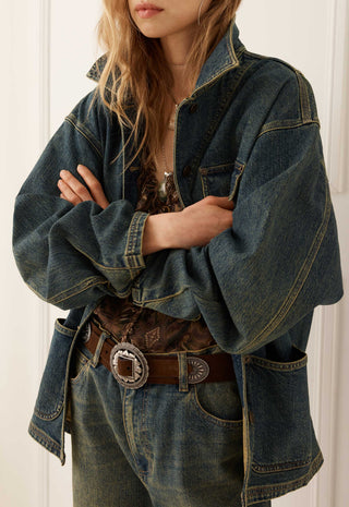 a-person-wearing-an-oversized-dark-blue-denim-jacket-with-wide-sleeves-paired-with-a-brown-patterned-top-a-brown-leather-belt-with-a-large-silver-concho-buckle-and-blue-jeans-set-against-a-white-paneled-background
