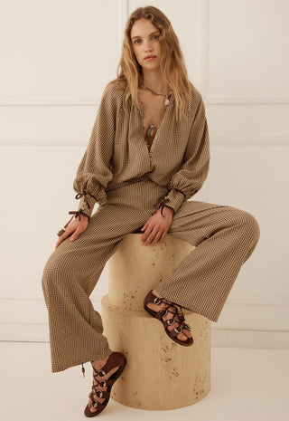 a-woman-sits-on-a-round-stone-pedestal-wearing-a-brown-and-beige-gingham-two-piece-set-consisting-of-a-long-sleeve-blouse-and-wide-leg-trousers-paired-with-brown-strappy-flat-sandals-against-a-white-paneled-wall