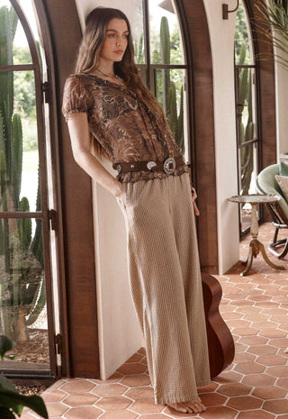 a-woman-stands-barefoot-in-a-rustic-indoor-setting-with-arched-windows-and-terracotta-tile-flooring-wearing-a-bohemian-style-outfit-consisting-of-a-brown-patterned-short-sleeve-top-a-wide-brown-belt-with-large-silver-conchos-and-beige-wide-leg-checkered-pants