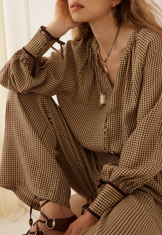 a-woman-wearing-a-brown-and-beige-gingham-long-sleeve-jumpsuit-with-tie-details-on-the-cuffs-paired-with-brown-strappy-sandals-and-silver-jewelry