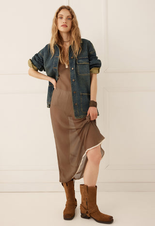 A woman models a bohemian-style outfit consisting of a brown midi slip dress, a blue denim jacket, and brown suede boots, set against a clean white paneled wall.