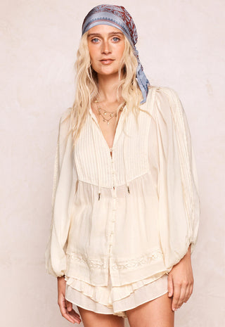 a-woman-wearing-a-cream-colored-long-sleeved-bohemian-style-blouse-with-pleated-details-and-a-ruffled-hem-paired-with-a-blue-patterned-headscarf-set-against-a-neutral-beige-background