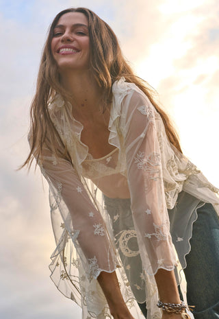a-woman-outdoors-wearing-a-sheer-cream-lace-bohemian-style-cover-up-over-a-matching-bralette-and-jeans-with-a-bright-sky-background
