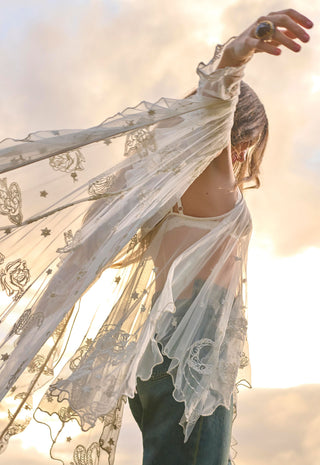 a-woman-outdoors-wearing-a-sheer-white-embroidered-lace-flutter-sleeve-cardigan-over-a-white-top-and-blue-jeans-with-her-arm-raised-against-a-bright-sunset-sky