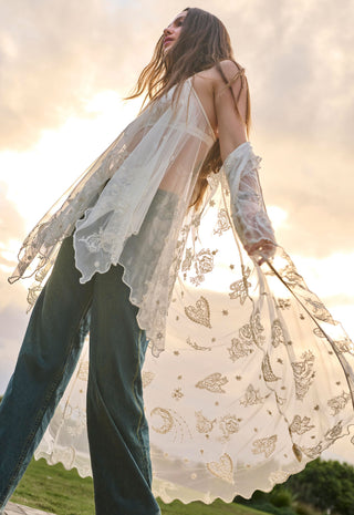 a-woman-outdoors-wearing-a-sheer-white-lace-long-sleeve-cardigan-with-gold-butterfly-and-moon-motifs-over-dark-teal-wide-leg-jeans-photographed-against-a-bright-sunset-sky