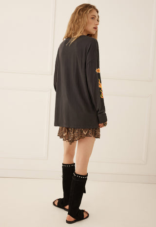 a-woman-is-shown-from-the-back-wearing-an-oversized-black-long-sleeve-top-with-orange-graphic-print-on-the-sleeve-a-brown-patterned-mini-skirt-and-black-knee-high-studded-boots-against-a-white-paneled-wall